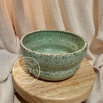A green ceramic tea bowl with a speckled pattern, placed on a wooden stand.
