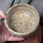 Hand holding a ceramic bowl with speckled glaze against a textured fabric background