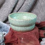 Speckled green ceramic bowl on a textured surface with fabric and stones.