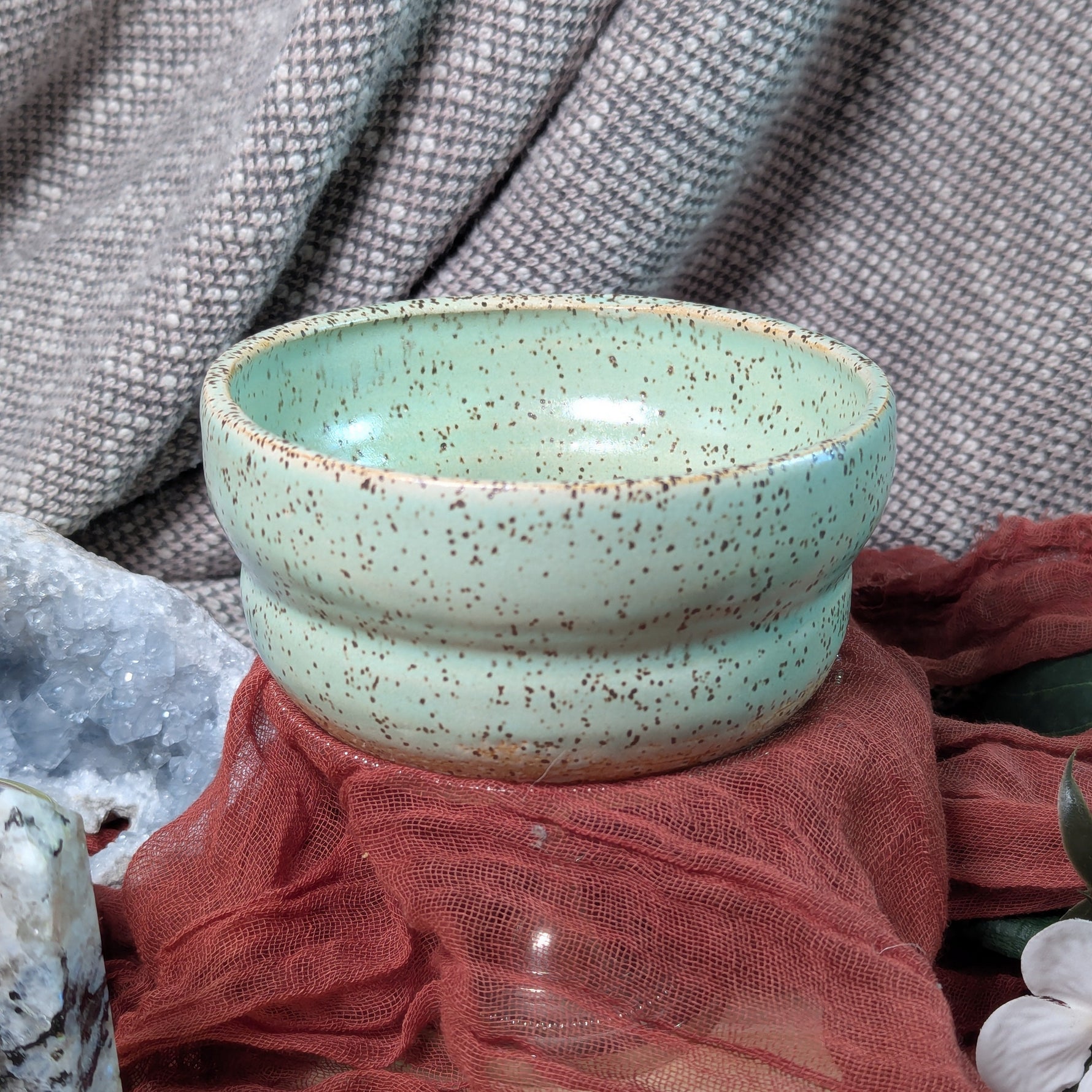 Speckled green ceramic bowl on a textured surface with fabric and stones.