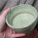 Hand holding a green ceramic bowl with speckled texture against a textured fabric background