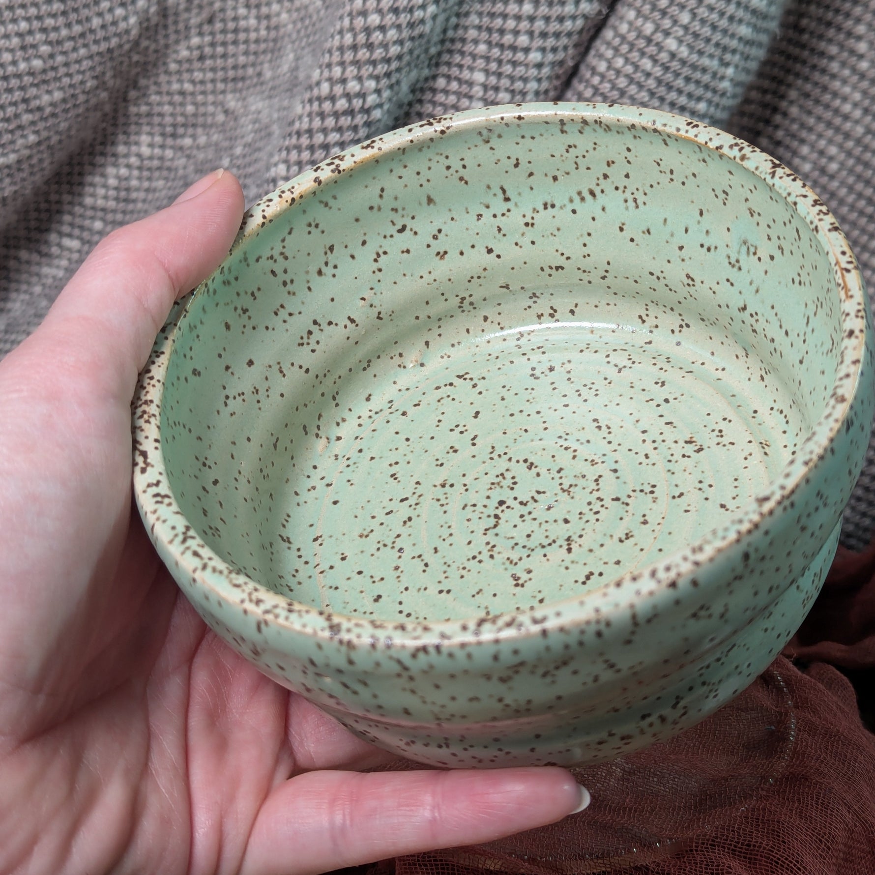 Hand holding a green ceramic bowl with speckled texture against a textured fabric background