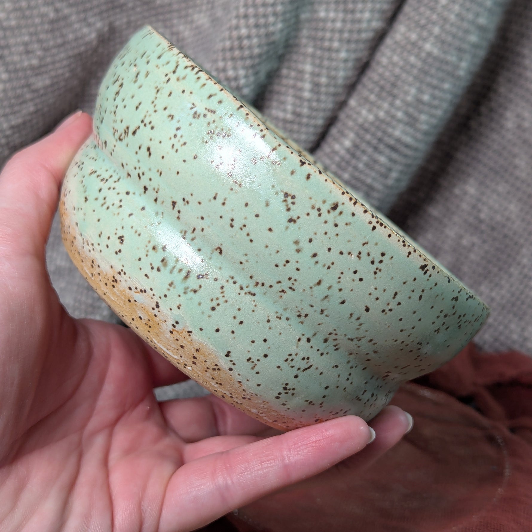 Hand holding a green ceramic bowl with speckled texture against a gray fabric background