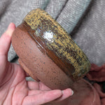 Hand holding a ceramic bowl with brown and yellow glaze against a textured gray background