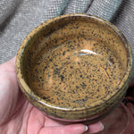 Hand holding a ceramic bowl with speckled design against a textured fabric background